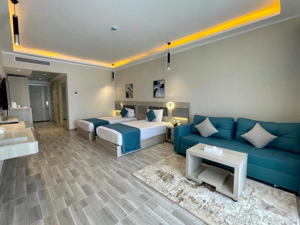 Moderna camera d'albergo con due letti singoli, biancheria da letto verde acqua e divano verde acqua con cuscini chiari. Il tavolino in legno chiaro e il tappeto neutro arricchiscono lo spazio. Le luci soffuse sul soffitto e l'arredamento minimalista evocano l'atmosfera invitante di Sirena Beach.