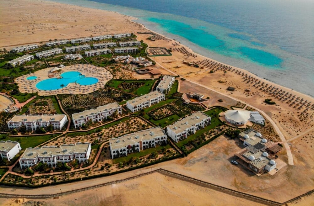 Vista aerea del Gorgonia Beach Resort con grandi piscine, edifici bianchi, giardini paesaggistici e una spiaggia fiancheggiata da ombrelloni, circondata dal deserto e affacciata su un mare turchese e trasparente.