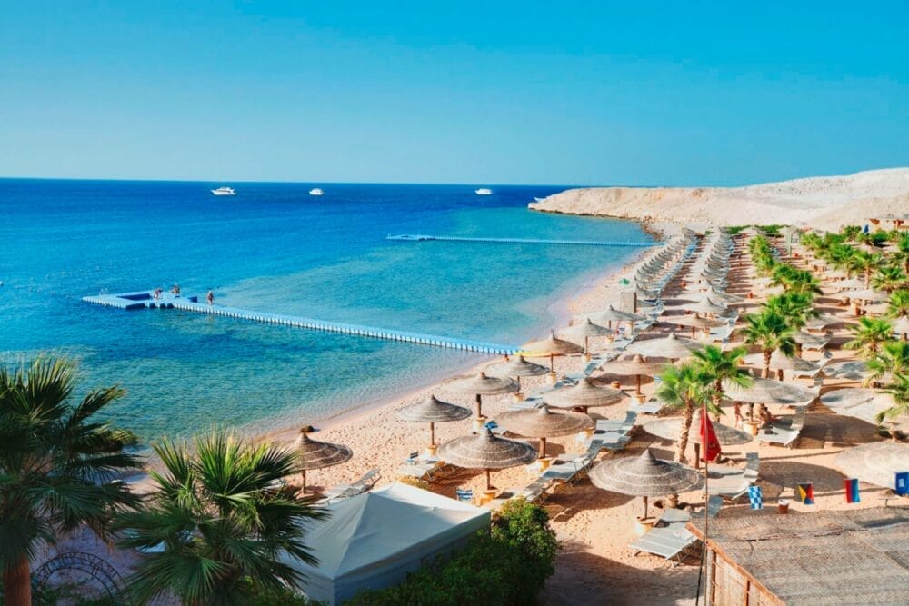 Una spiaggia sabbiosa a Sharm El Sheikh con file di ombrelloni di paglia e sedie a sdraio, palme e un lungo molo che si protende nell'acqua azzurra e trasparente. Le barche galleggiano in lontananza sotto un cielo luminoso e soleggiato vicino al Seaclub Savoy.