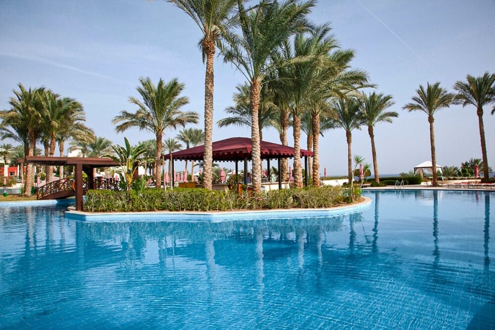 Una grande piscina all'aperto con acqua cristallina al Grand Rotana Resort, circondata da palme e da un padiglione con tetto rosso al centro, sotto un cielo limpido: l'ideale per rilassarsi al Seaclub o godersi la spa.