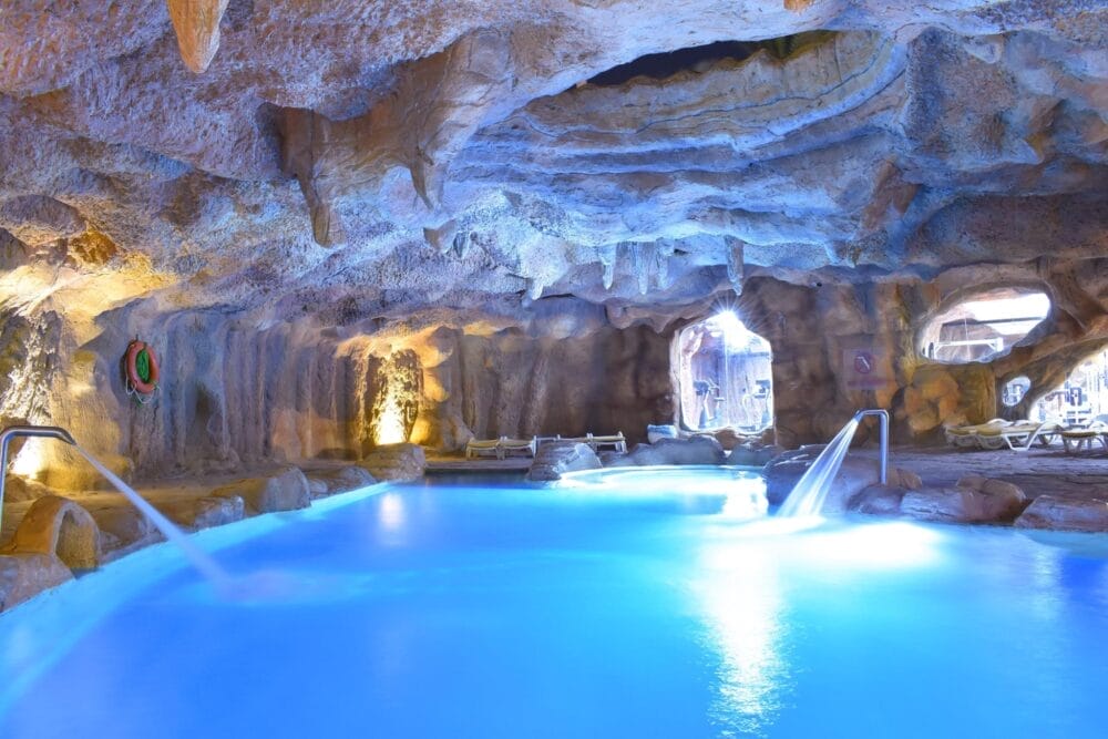 Una piscina interna luminosa all'interno di una stanza simile a una grotta con pareti e soffitto di roccia al Parrotel Beach Resort, con sedie a sdraio, flussi d'acqua che confluiscono nella piscina e luci blu soffuse: un'oasi che ricorda l'atmosfera del resort sulla spiaggia di Sharm El Sheikh.