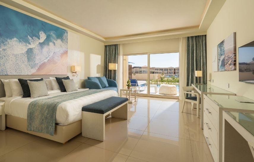 Moderna camera d'albergo del Cleopatra Luxury Resort con letto grande, opere d'arte a tema oceanico, decorazioni blu e bianche, porte di vetro che si aprono su un balcone con vista sulla piscina e un'area salotto vicino alla finestra.