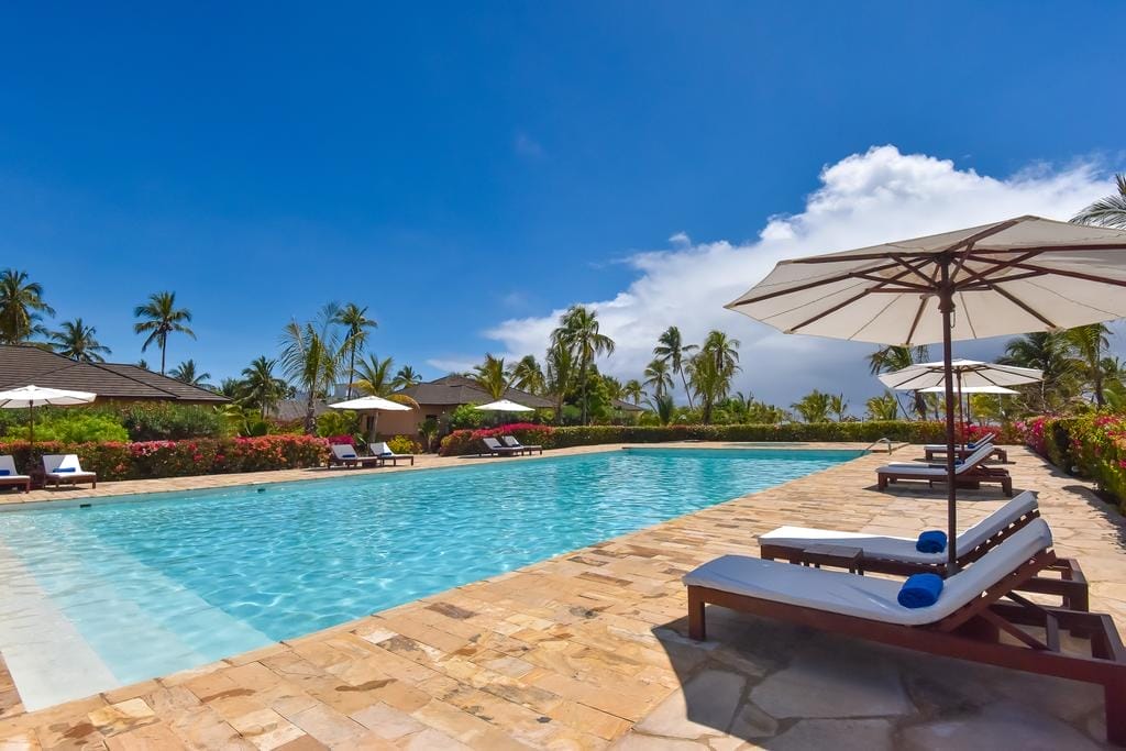 Una lussuosa piscina all'aperto, dotata di Bozza automatica, è circondata da sedie a sdraio con ombrelloni bianchi, da una vegetazione lussureggiante, da alte palme e da un cielo blu brillante punteggiato di nuvole.