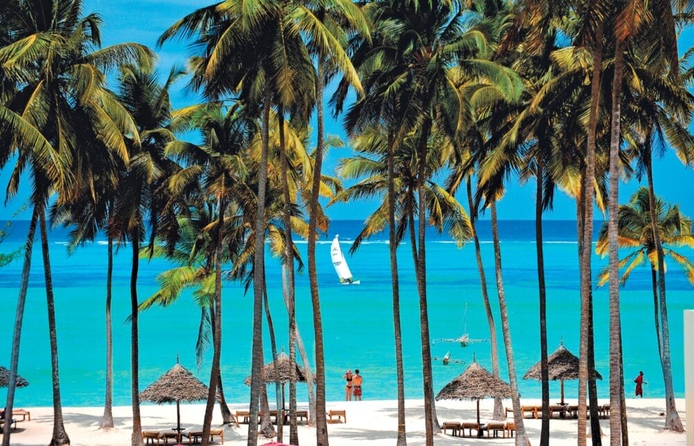 Alte palme costeggiano una spiaggia tropicale da sogno con sabbia bianca, lettini in legno e ombrelloni di paglia, in puro stile Seaclub. L'oceano turchese brilla alle spalle, con una barca a vela alla deriva e persone che passeggiano sulla riva.