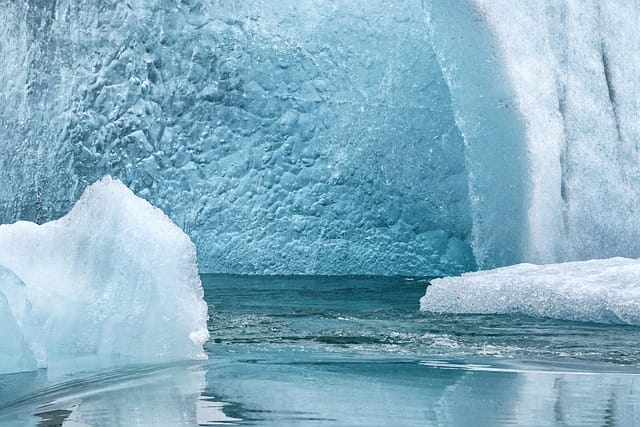 Grandi iceberg azzurri e strutturati emergono dall'acqua fredda e limpida; le loro superfici presentano crepe e creste, con riflessi visibili sull'acqua ferma sottostante: una scena che ricorda un dipinto automatico di Bozza.
