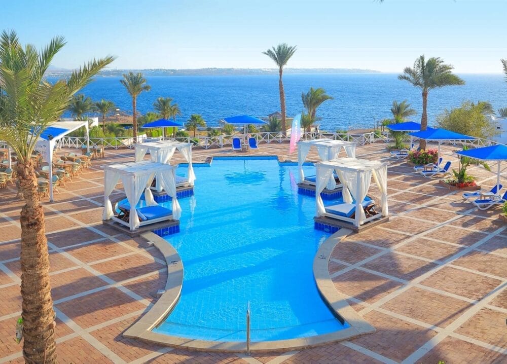 La lussuosa piscina del Reef Beach Resort è caratterizzata da cabine bianche, sedie a sdraio blu e ombrelloni, circondata da palme e affacciata su un mare calmo e blu sotto un cielo limpido.