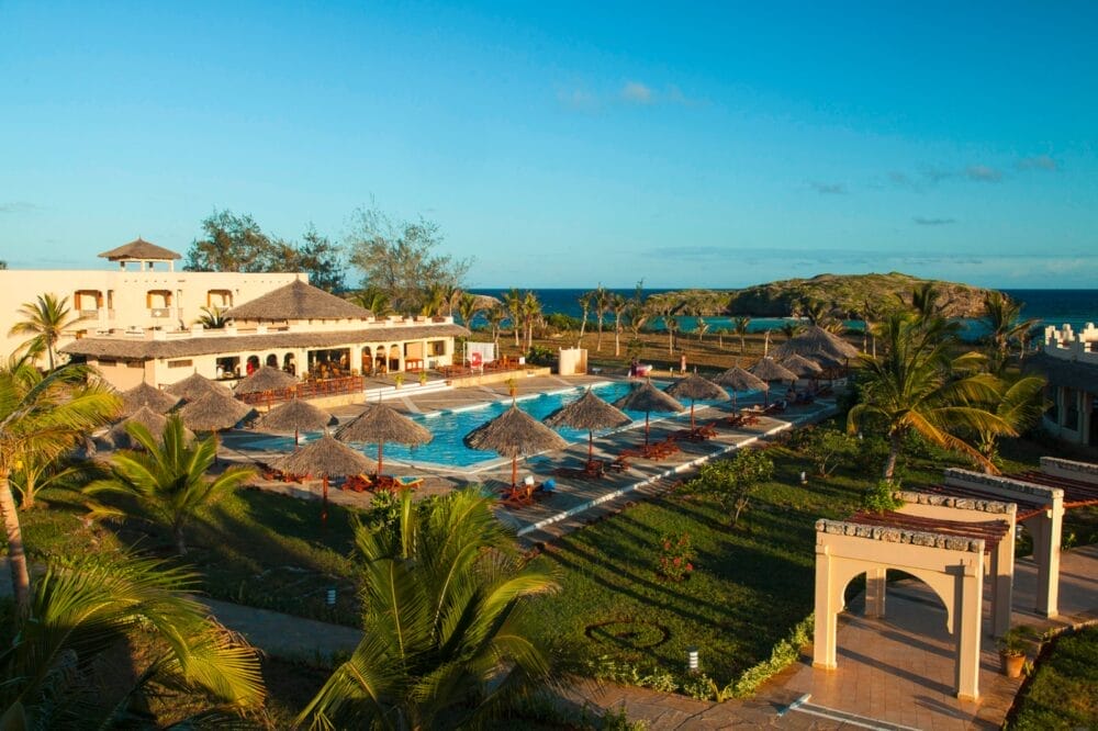 Uno splendido Island Resort con una grande piscina all'aperto, lettini e ombrelloni di paglia, circondato da palme e giardini, che si affaccia sull'oceano sotto un cielo azzurro e limpido.