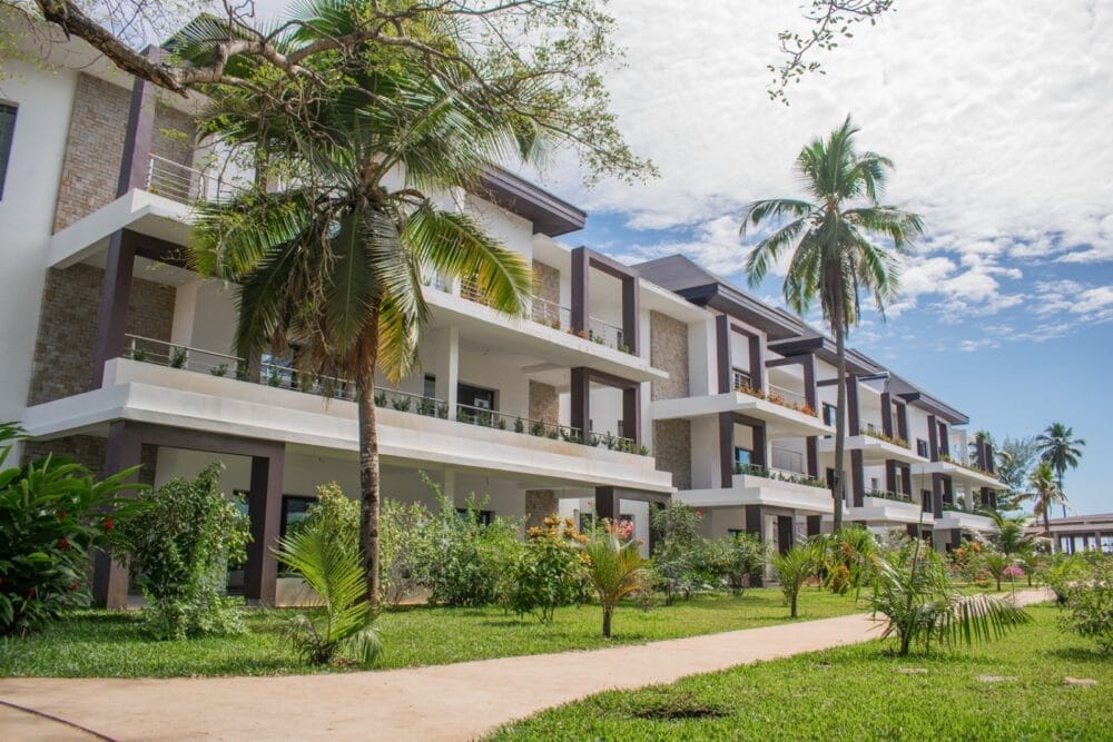 Moderno edificio alberghiero a tre piani con balconi, circondato da piante tropicali e palme, sotto un cielo parzialmente nuvoloso. Una passerella pavimentata corre lungo il prato verde di fronte al Seaclub Royal Andilana Resort and Spa.
