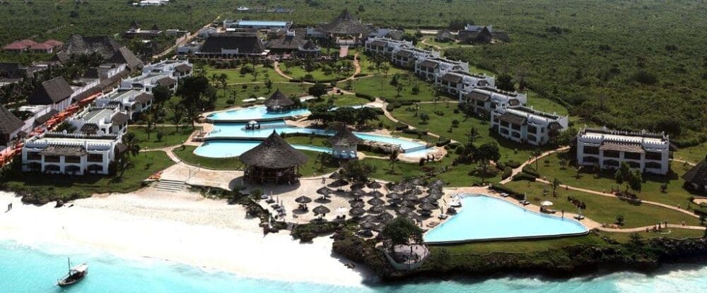 Vista aerea del Royal Zanzibar Beach Resort con diverse piscine, edifici bianchi, strutture con tetti di paglia e lettini su una spiaggia sabbiosa accanto all'acqua turchese dell'oceano, circondata da una vegetazione lussureggiante.