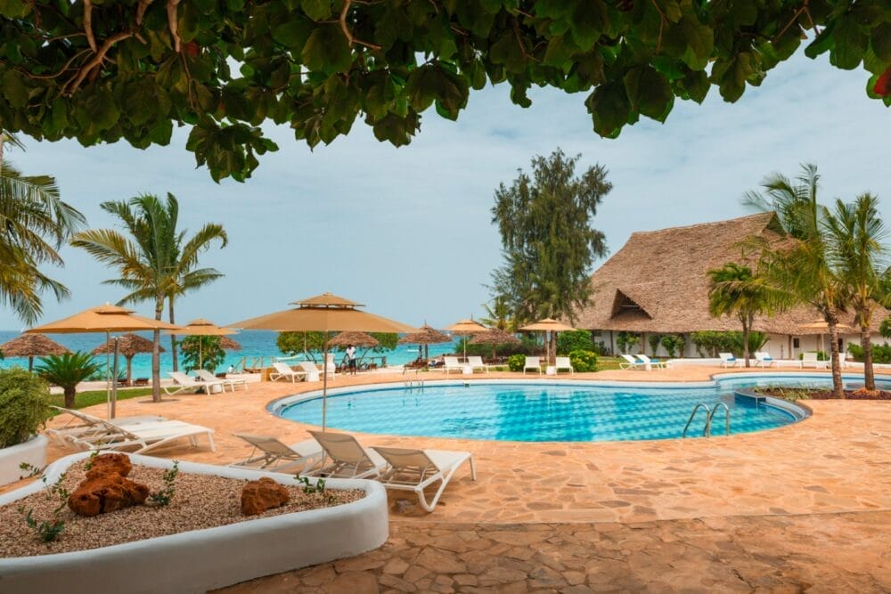 Il resort tropicale Seaclub è caratterizzato da una piscina rotonda circondata da sedie a sdraio e ombrelloni, con palme, verde lussureggiante e un edificio con tetto di paglia vicino all'oceano blu sotto un cielo parzialmente nuvoloso.