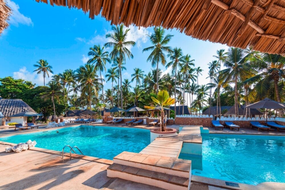 La piscina del resort tropicale Seaclub Diamond Mapenzi Beach, circondata da palme, lettini e ombrelloni sotto un cielo blu brillante; un tetto di paglia copre parzialmente il primo piano.
