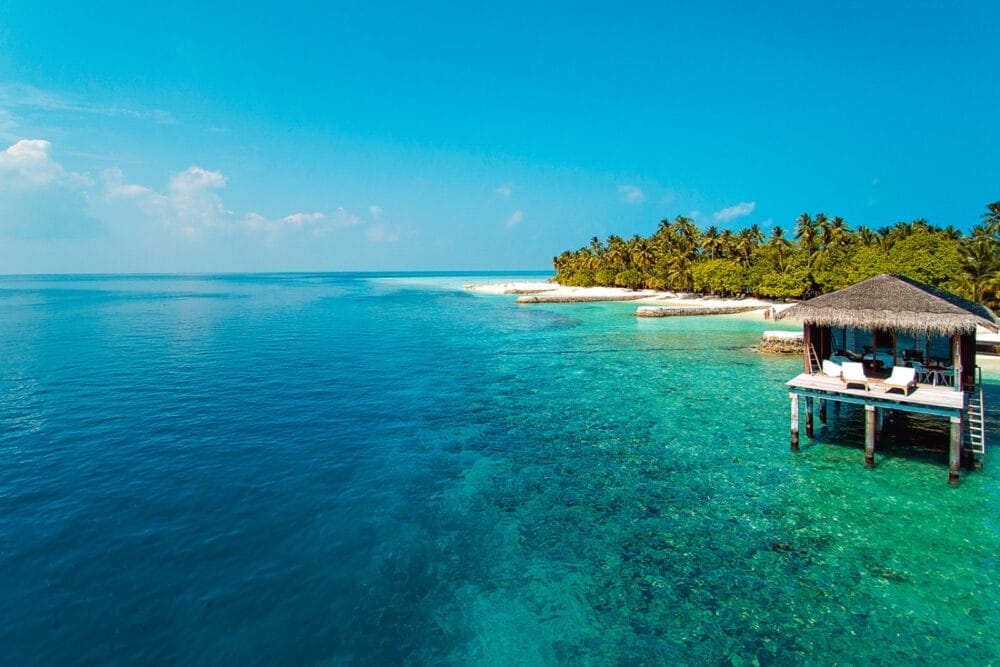 Un'isola tropicale con palme lussureggianti, spiagge di sabbia bianca e acque turchesi. Una cabana in legno del resort Seaclub Dhiggiri Maldives si affaccia sull'oceano limpido, di fronte al mare aperto sotto un cielo blu brillante.