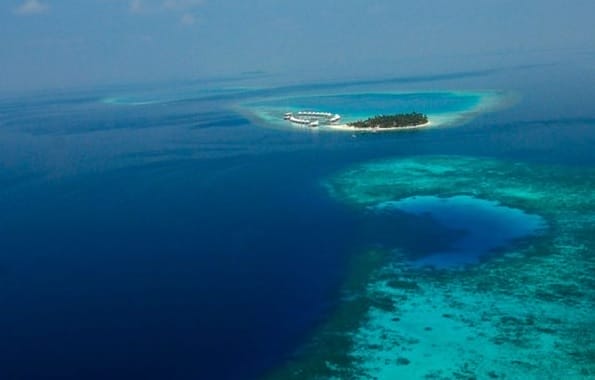 Vista aerea di una piccola isola con Water Villas ed edifici, circondata da acque turchesi, barriere coralline e oceano blu intenso alle Maldive.