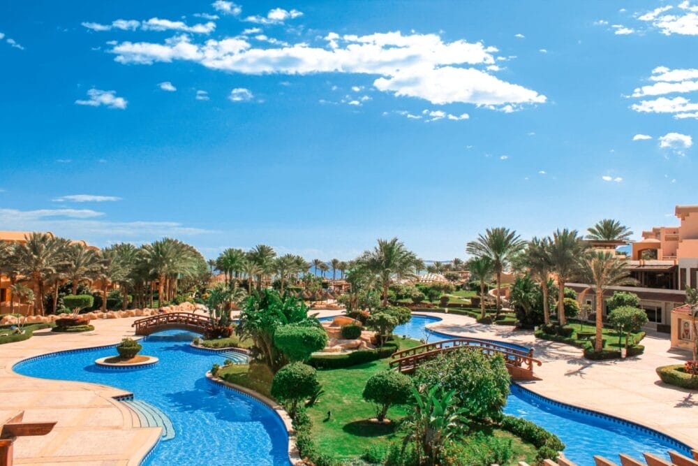 Scoprite il Grand Plaza Resort, una fuga di lusso con piscine blu, ponti di legno ad arco, giardini lussureggianti e palme ondeggianti sotto un cielo azzurro punteggiato di nuvole.