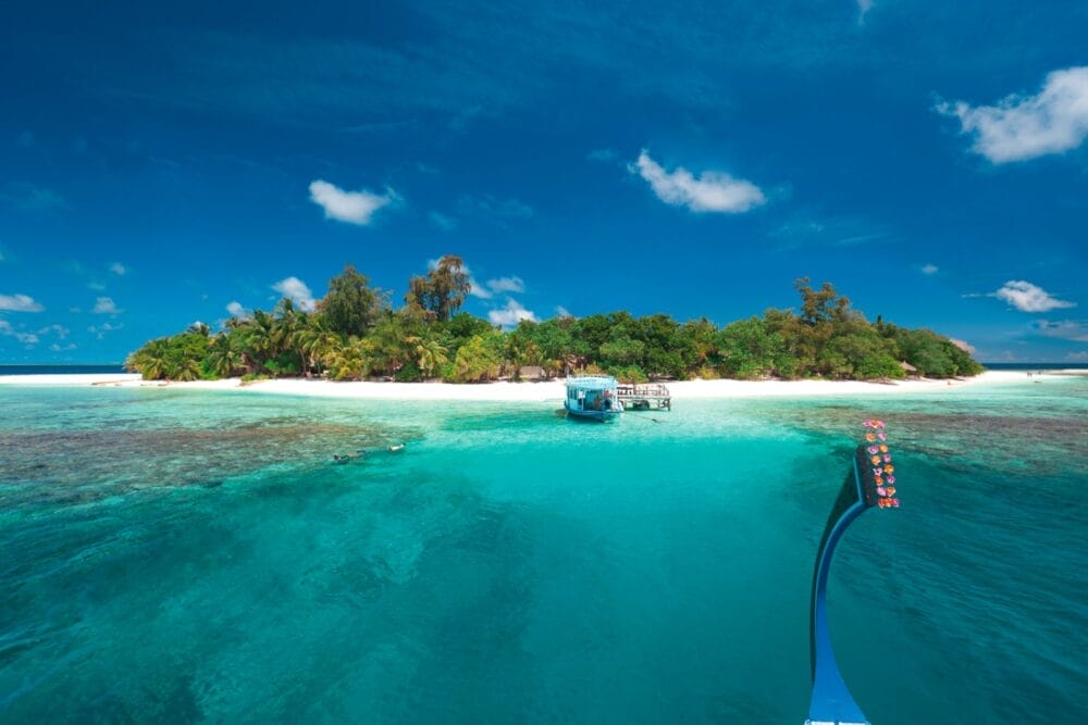 Una piccola isola tropicale con alberi verdi e una spiaggia di sabbia bianca è circondata da acque limpide e turchesi vicino al Sandies Bathala Resort. Una barca è ancorata a riva, con un'altra barca parzialmente visibile in primo piano sotto un cielo blu brillante.