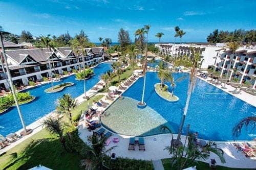 Vista aerea del Sunprime Kamala Beach Resort con diverse piscine, palme, sedie a sdraio ed edifici bianchi sotto un cielo azzurro e limpido, con una vegetazione lussureggiante e l'oceano vicino alla spiaggia di Kamala sullo sfondo.