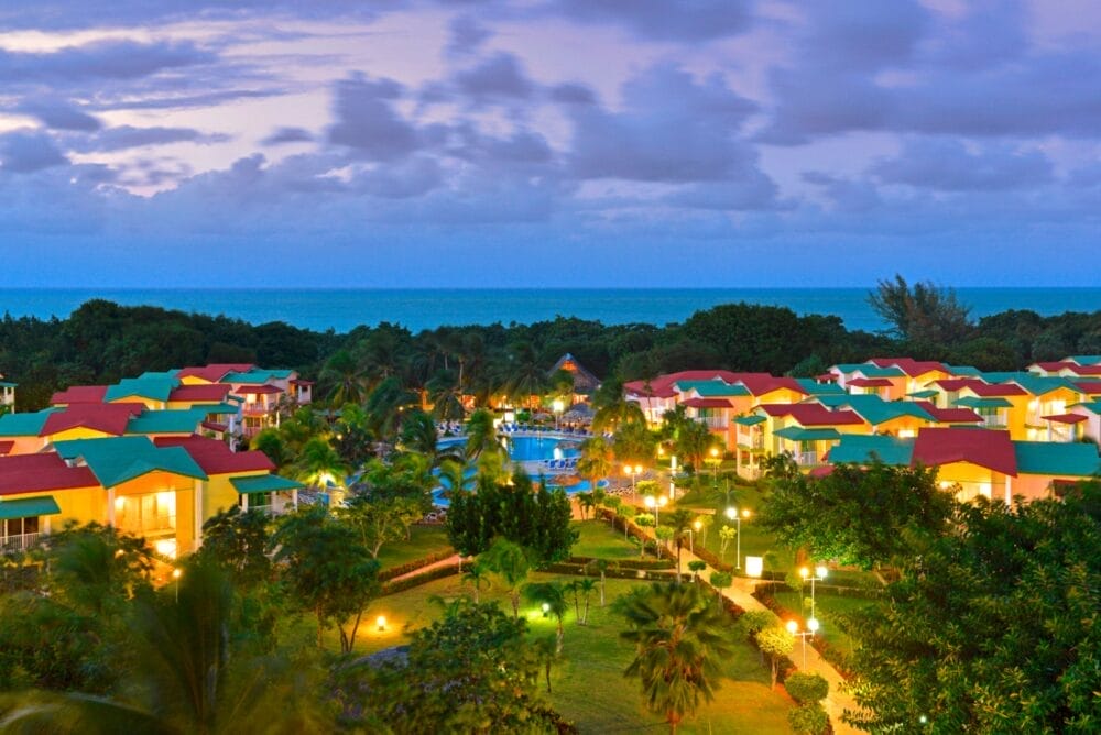 Vista aerea del resort Seaclub Iberostar Tainos al tramonto, con i tetti colorati di rosso, verde e giallo incastonati tra palme e giardini. L'oceano brilla sullo sfondo sotto il cielo nuvoloso della sera.