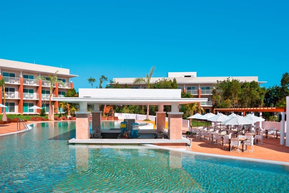 Al resort Playa Vista Azul vi attende una luminosa piscina all'aperto con bar, circondata da sedie a sdraio e ombrelloni. Sullo sfondo, sotto un cielo azzurro e limpido, sorgono moderni edifici alberghieri con balconi.