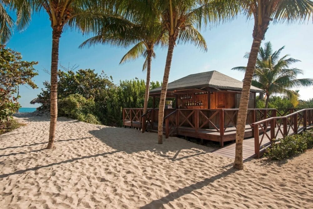 Un gazebo in legno sulla spiaggia del Searesort è circondato da palme su un terreno sabbioso, con la luce del sole che proietta ombre e verde lussureggiante sullo sfondo sotto un cielo azzurro e limpido.