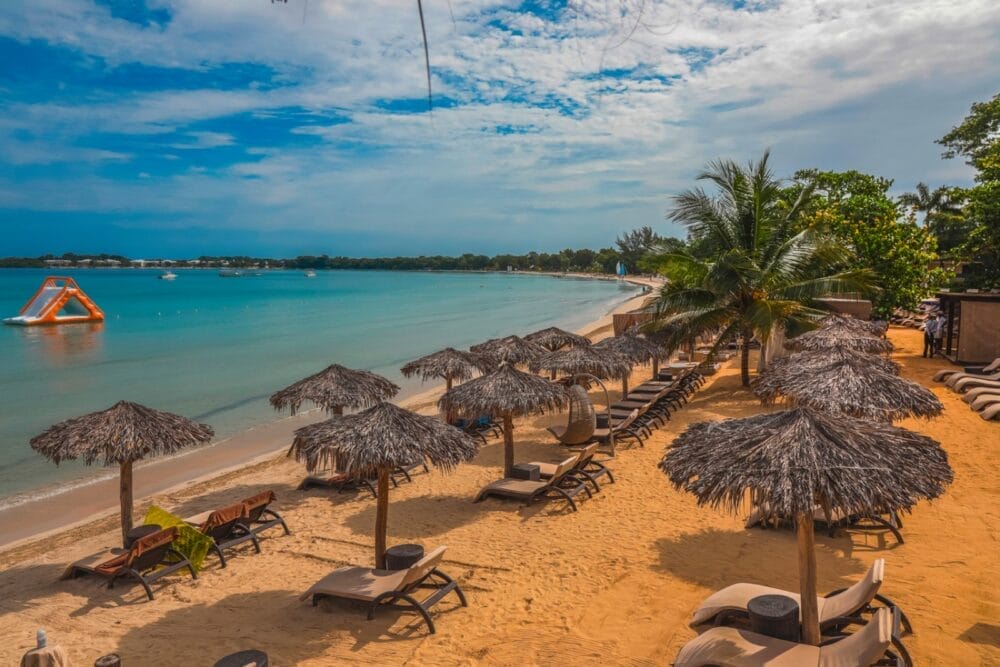 Una spiaggia tropicale al Royalton Negril con file di sedie a sdraio vuote sotto ombrelloni di paglia sulla sabbia dorata che si affaccia su un mare azzurro e calmo. Le palme costeggiano la riva e una struttura gonfiabile arancione galleggia in mare sotto un cielo parzialmente nuvoloso.