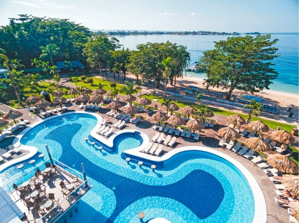 Vista aerea della piscina del resort Riu Negril dalle forme curve, circondata da lettini e ombrelloni di paglia, con palme e prati verdi nelle vicinanze, accanto alla spiaggia sabbiosa di Negril e al mare azzurro e calmo sotto un cielo limpido.