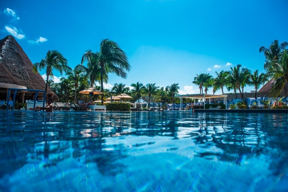 Una luminosa giornata di sole nella piscina del resort Catalonia Playa Maroma, con acqua cristallina, palme, sedie a sdraio, ombrelloni ed edifici con tetti di paglia. Il cielo è limpido e le persone si rilassano a bordo piscina.