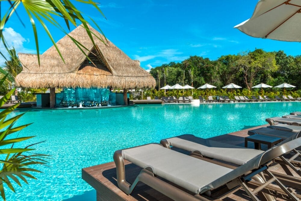 Le sedie a sdraio costeggiano la grande piscina all'aperto dell'Ocean Riviera Paradise, con un bar sul tetto di paglia sullo sfondo, circondato da palme e ombrelloni bianchi sotto il cielo azzurro di questo splendido resort sul mare.