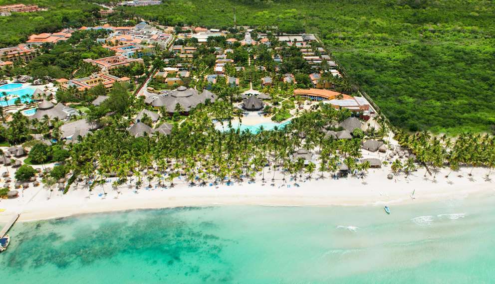 Vista aerea del Seaclub Catalonia Bayahibe, un resort tropicale con piscine, palme e vegetazione lussureggiante, delimitato da una spiaggia di sabbia bianca e dalle acque turchesi dell'oceano a Bayahibe. Lungo il litorale sono visibili ombrelloni e persone.