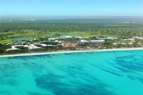Veduta aerea del Barcelo Bavaro Palace di Punta Cana, un resort all-inclusive con acque turchesi e trasparenti, un litorale di sabbia bianca e una vegetazione lussureggiante che si estende nell'entroterra sotto un cielo blu brillante.