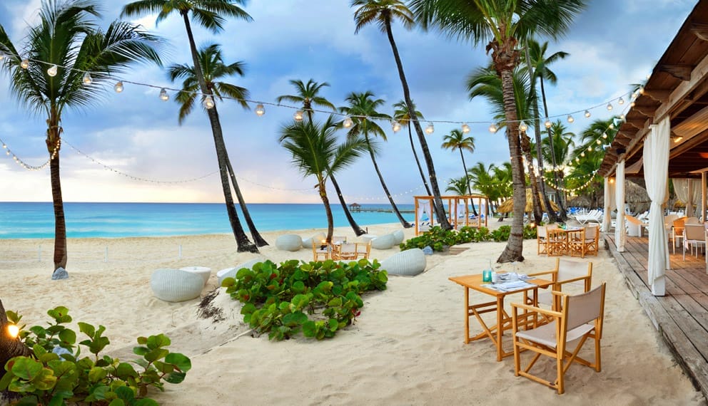 Cena all'aperto sulla spiaggia del Searesort, Catalonia Royal La Romana, un'oasi di oltre 18 tavoli in legno sulla riva sabbiosa, circondata da palme e luci, con vista sull'oceano turchese sotto un cielo parzialmente nuvoloso.