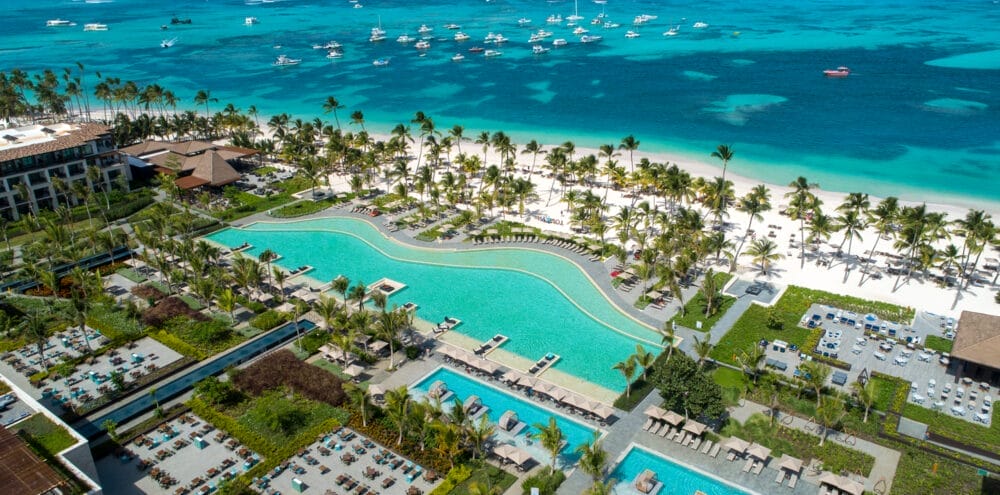 Vista aerea del Lopesan Costa Bavaro, un resort di lusso sulla spiaggia con grandi piscine, sedie a sdraio, palme e una spiaggia di sabbia bianca accanto a un oceano limpido e turchese, con barche ancorate al largo.