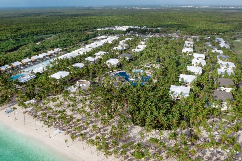 Vista aerea del resort Catalonia Bavaro Beach, con edifici bianchi circondati da palme verdi e lussureggianti, diverse piscine e un litorale sabbioso accanto all'acqua turchese dell'oceano, che crea un'atmosfera vibrante da club marino.