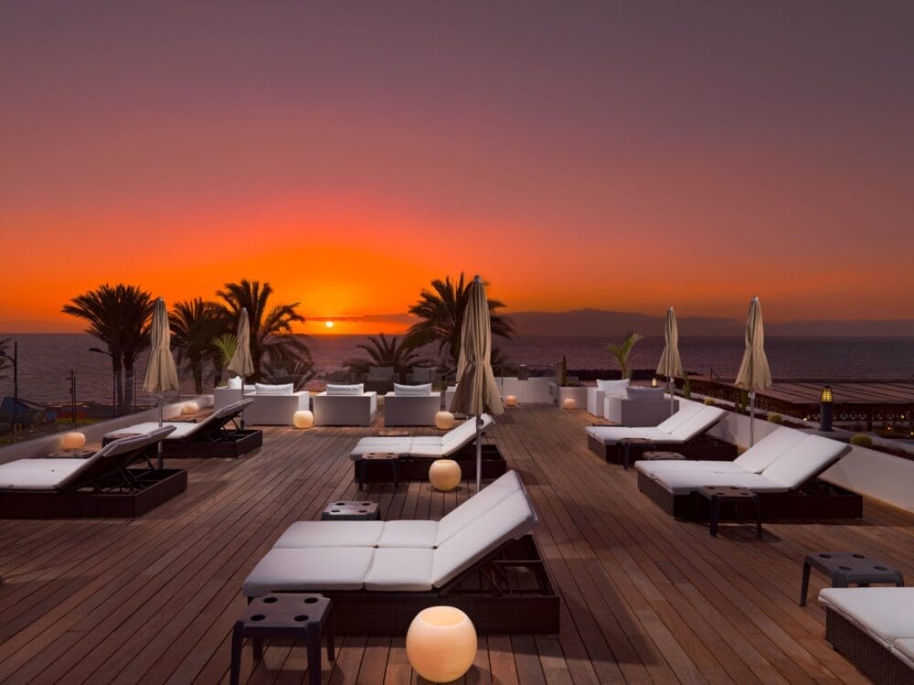 La terrazza sul tetto del Seaclub H10 Gran Tinerfe è dotata di moderne sedie a sdraio e ombrelloni con vista sull'oceano al tramonto. Le palme incorniciano la vista, mentre le luci rotonde e scintillanti creano un'atmosfera rilassante sul ponte di legno.