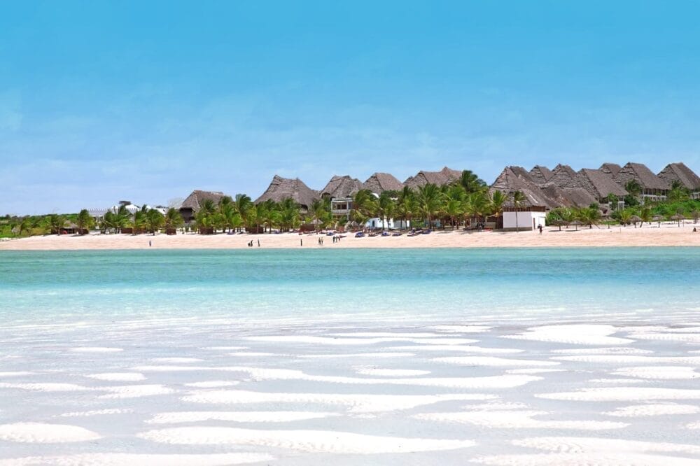 Spiaggia di sabbia bianca con acqua limpida e turchese in primo piano, alcuni edifici con tetti di paglia e palme sul retro e un cielo azzurro brillante in alto. Alcune persone passeggiano lungo il litorale di Bravo Watamu.