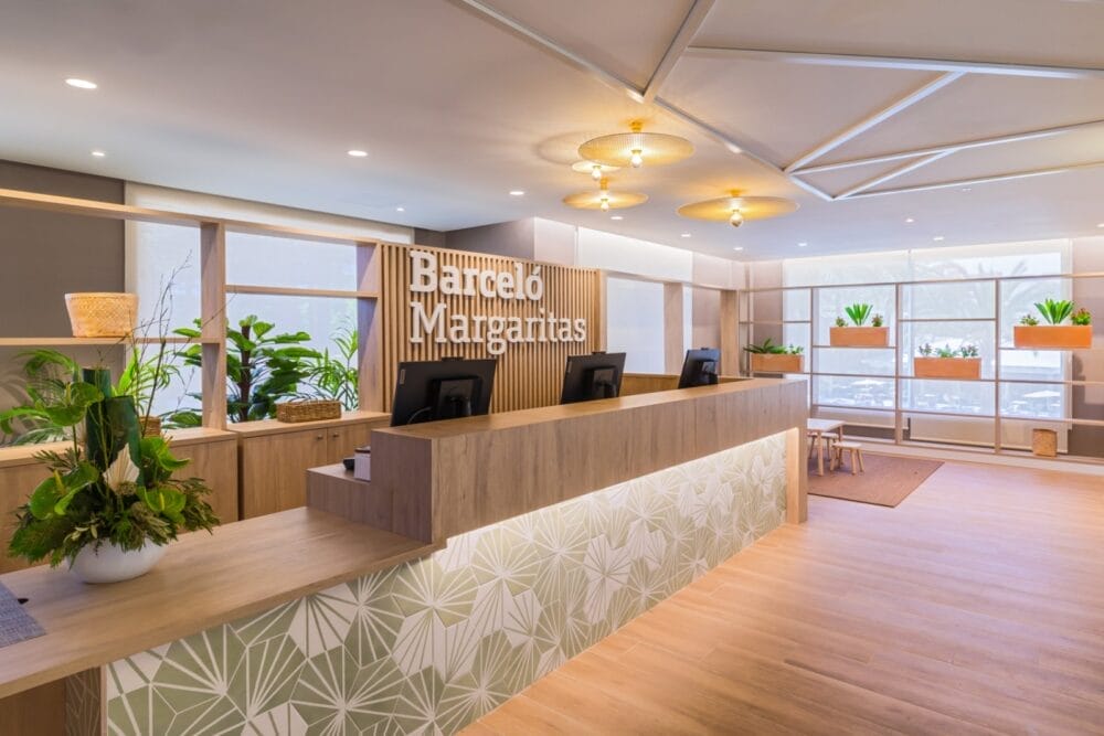 Moderna area reception dell'hotel con mobili in legno chiaro, piante verdi e grandi finestre. La reception presenta motivi geometrici e un'insegna con la scritta "Seaclub Barcelò Margaritas". Lo spazio è luminoso e accogliente.