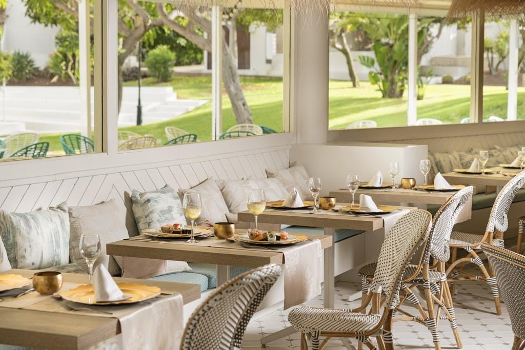 Il ristorante dell'Alua Suites Fuerteventura è luminoso ed elegante, con tavoli in legno, panche imbottite e sedie in vimini. Le ampie finestre si affacciano su un giardino verde e paesaggistico, mentre il bianco e i toni neutri creano un'atmosfera rilassata e ariosa da resort marittimo.