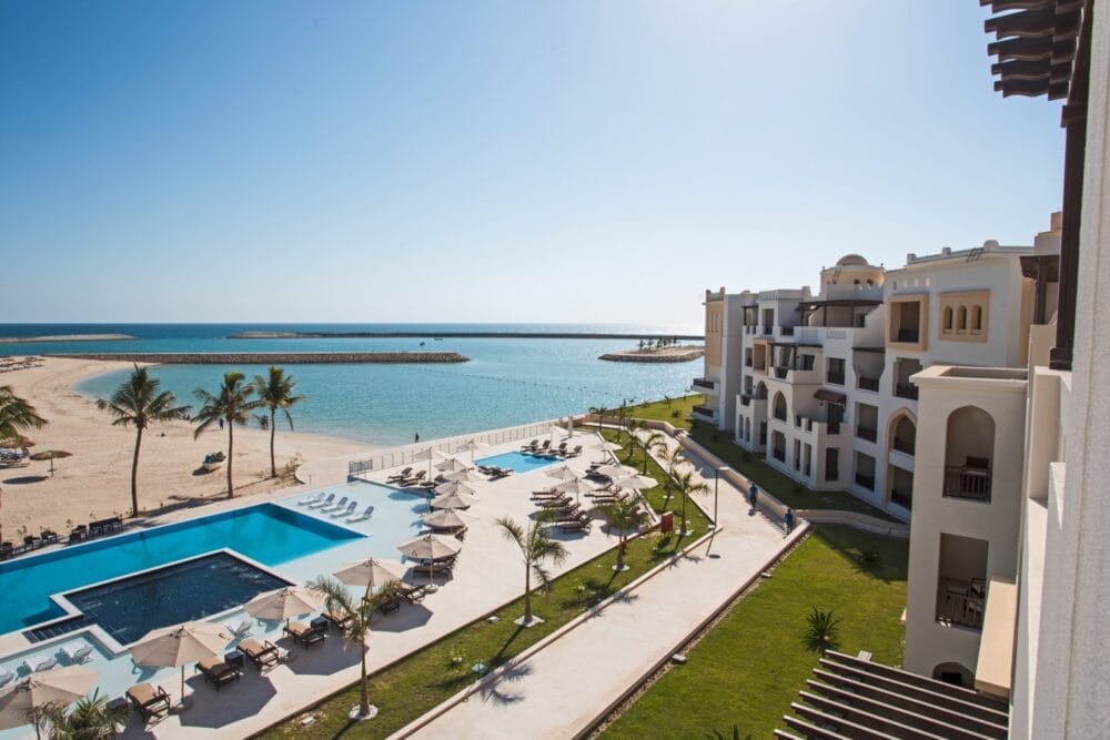 Il Seaclub Fanar è un resort sulla spiaggia con un'ampia piscina, lettini e ombrelloni che si affacciano su una spiaggia sabbiosa e un mare azzurro e calmo. Moderni edifici bianchi con balconi si affacciano sulla piscina e sul litorale sotto un cielo limpido e soleggiato.