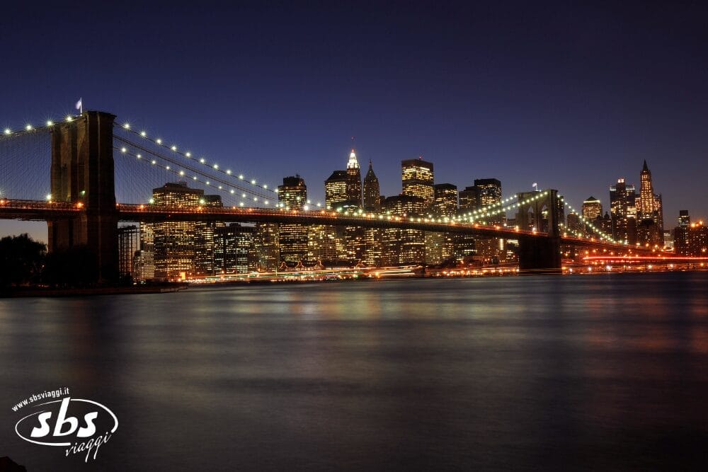 Il ponte di Brooklyn illuminato di notte attraversa l'acqua, con lo skyline di New York che si staglia sullo sfondo sotto un cielo blu intenso: una scena perfetta per un'acquisizione automatica di Bozza.