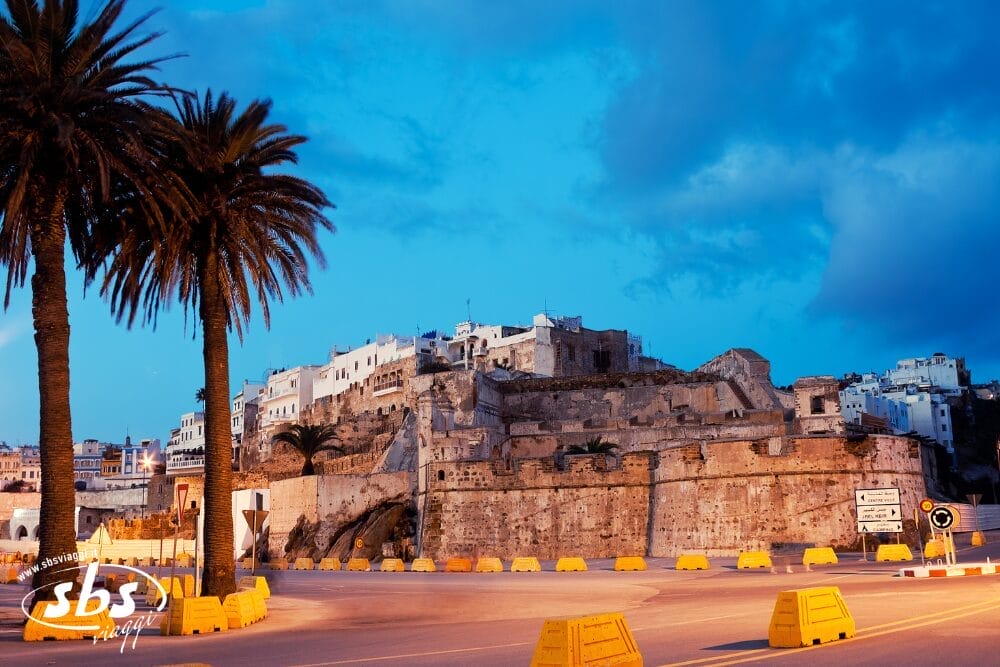 Una veduta di una fortezza storica con edifici bianchi su una collina al crepuscolo, circondata da palme e barriere gialle lungo la strada in primo piano: un'affascinante Bozza automatica sotto un cielo azzurro e carico di nuvole.