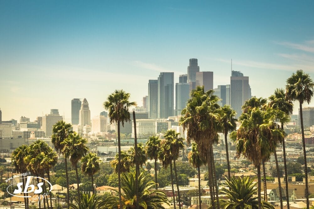 Lo skyline del centro di Los Angeles con alti edifici per uffici e file di palme in primo piano in una limpida giornata di sole. Il logo SBS e "Bozza automatica" sono visibili nell'angolo in basso a sinistra.