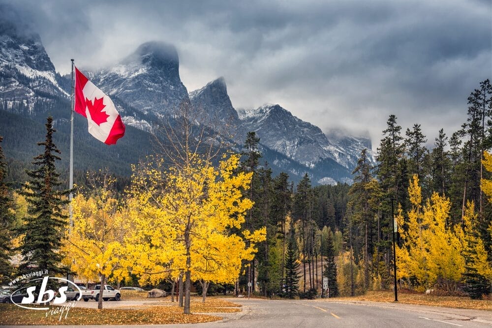Una bandiera canadese sventola accanto a vibranti alberi autunnali gialli, con alti pini e drammatiche Montagne Rocciose coperte di nuvole sullo sfondo, sotto un cielo lunatico: un vero Bozza automatico della bellezza stagionale della natura.