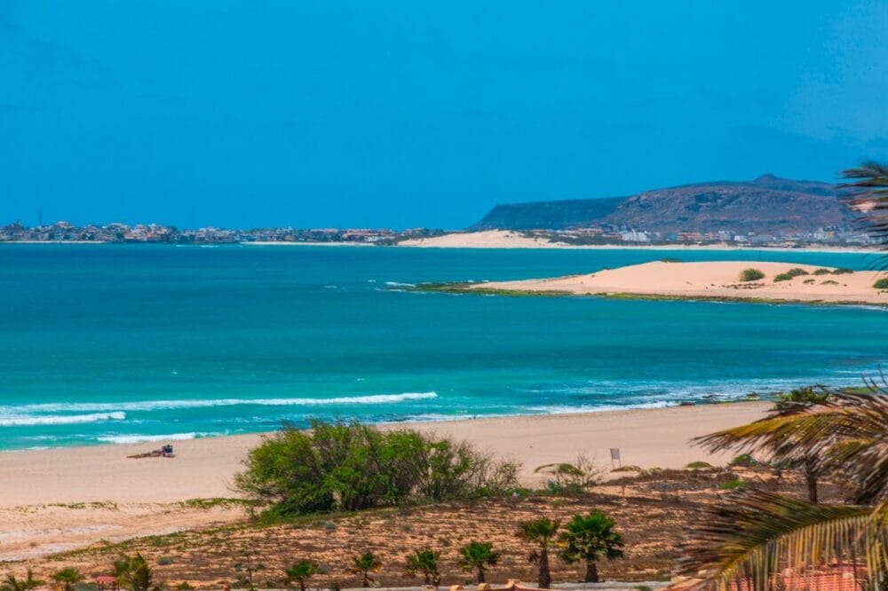 Un vivace paesaggio costiero caratterizzato da acque turchesi, spiagge sabbiose, vegetazione rada e palme in primo piano, con colline lontane sotto un cielo blu brillante, tipico dei panorami mozzafiato di Boa Vista Beach.