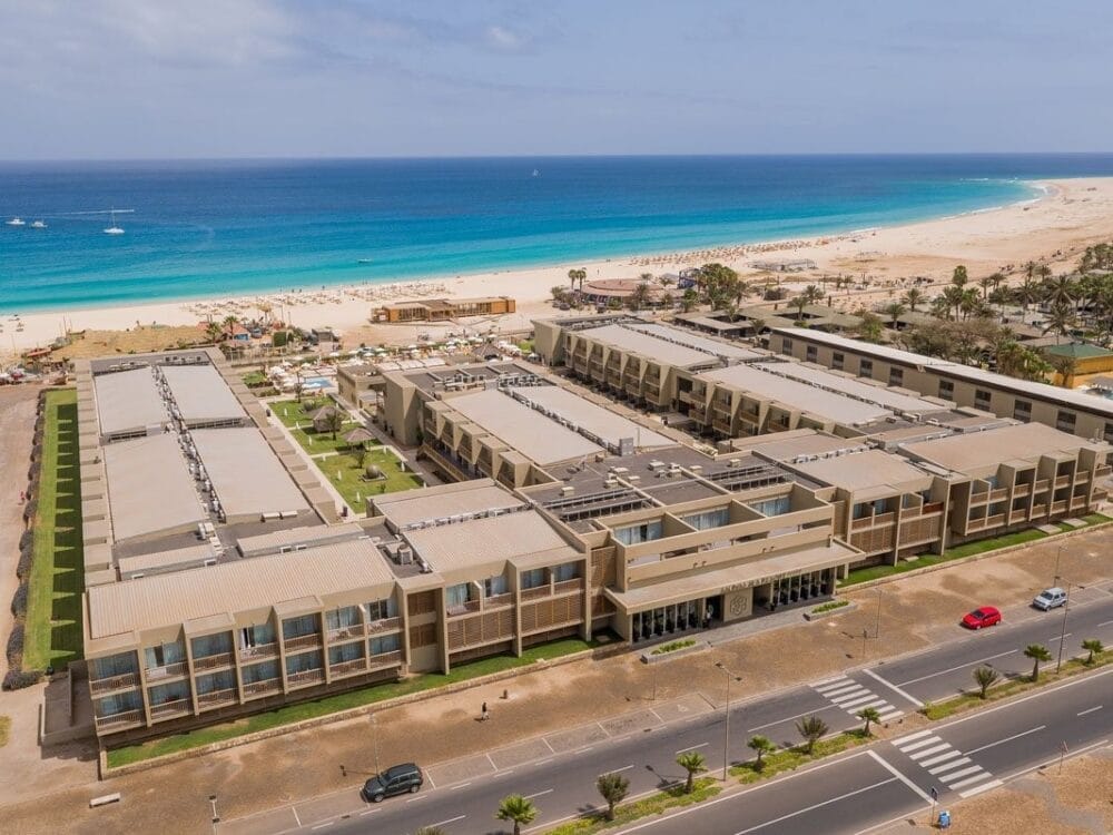 Vista aerea del resort Oasis Salinas Sea, con diversi edifici rettangolari, prati verdi e piscine, accanto a una spiaggia sabbiosa e all'oceano turchese sotto un cielo limpido. Una strada con automobili passa davanti a questa splendida destinazione Veraclub.