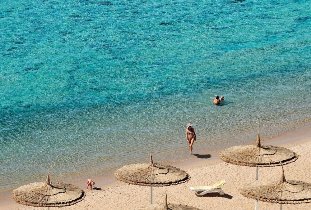 Un mare limpido e turchese e una spiaggia sabbiosa con ombrelloni di paglia al Reef Oasis Senses, una persona che cammina sulla riva, due persone che nuotano e una sedia a sdraio che si crogiola sotto il sole.