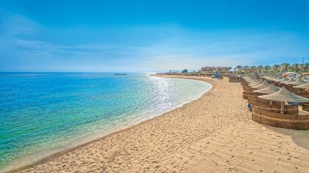 Una spiaggia sabbiosa con file di ombrelloni e sedie a sdraio sulla destra, un mare azzurro e calmo sulla sinistra e un cielo limpido in alto. Le palme e gli edifici del resort Veraclub Utopia Beach sono visibili in lontananza.