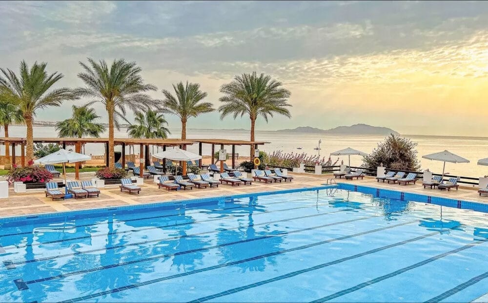 La grande piscina all'aperto del Veraclub Sharm è circondata da sedie a sdraio, ombrelloni e palme, e si affaccia su un mare calmo all'alba o al tramonto, con le montagne in lontananza: una fuga perfetta dal resort di Sharm el Sheikh.