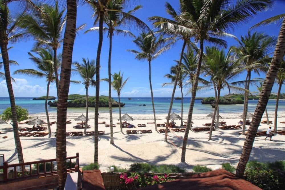 Vista di una spiaggia tropicale del resort Veraclub Crystal Bay, con alte palme, sabbia bianca, lettini e ombrelloni di paglia. L'oceano blu e le piccole isole verdi brillano sotto un cielo limpido.