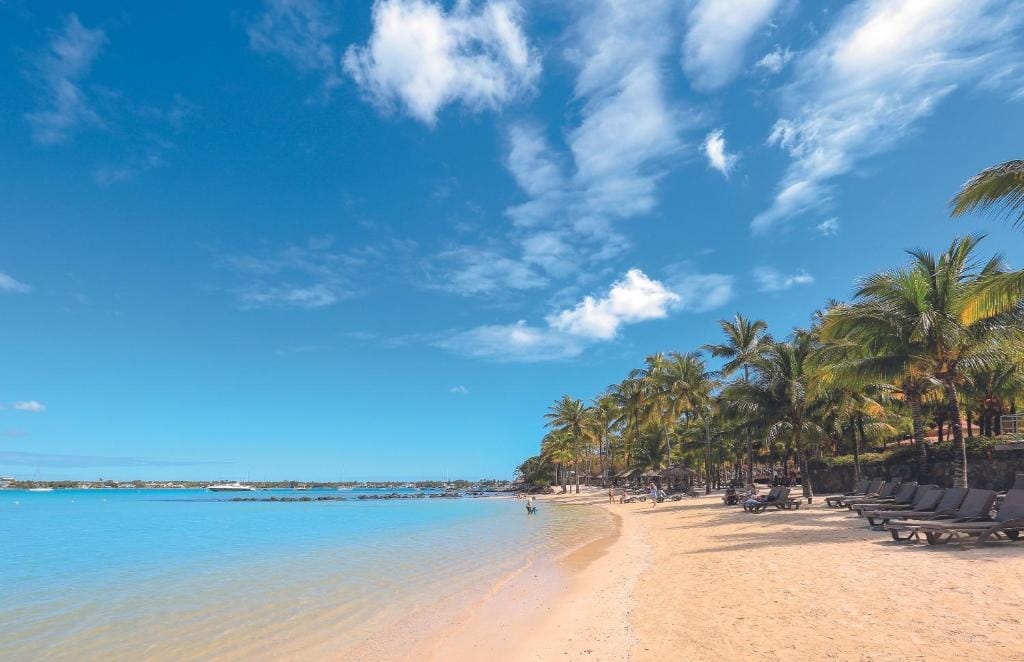 Una soleggiata spiaggia tropicale con sabbia dorata, acqua cristallina e palme ondeggianti al Mauricia Beachcomber, dove sedie a sdraio vuote attendono sotto un cielo blu con nuvole sparse.
