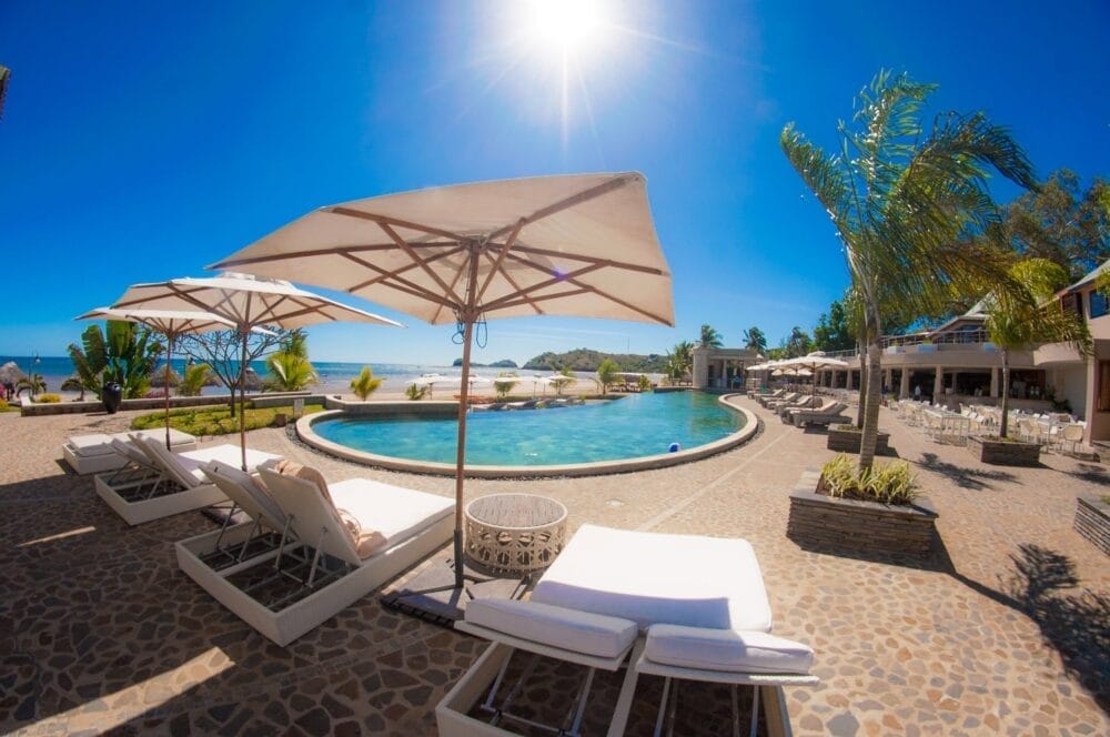 Scena di sole a bordo piscina del resort Veraclub Palm Beach, con sedie a sdraio bianche sotto grandi ombrelloni, palme ondeggianti, un ponte piastrellato in pietra e una piscina rotonda sotto un cielo azzurro e limpido.