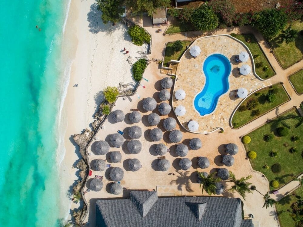 Vista aerea del resort Veraclub Sunset Beach con ombrelloni di paglia sulla sabbia, acqua turchese dell'oceano, una piscina sinuosa e un paesaggio verde che circonda l'area della piscina.